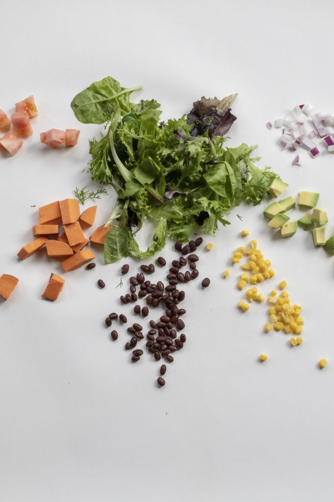 Salad surrounded by diced tomatoes, sweet potatoes, black beans, corn, avocado, and red onions on a white background.