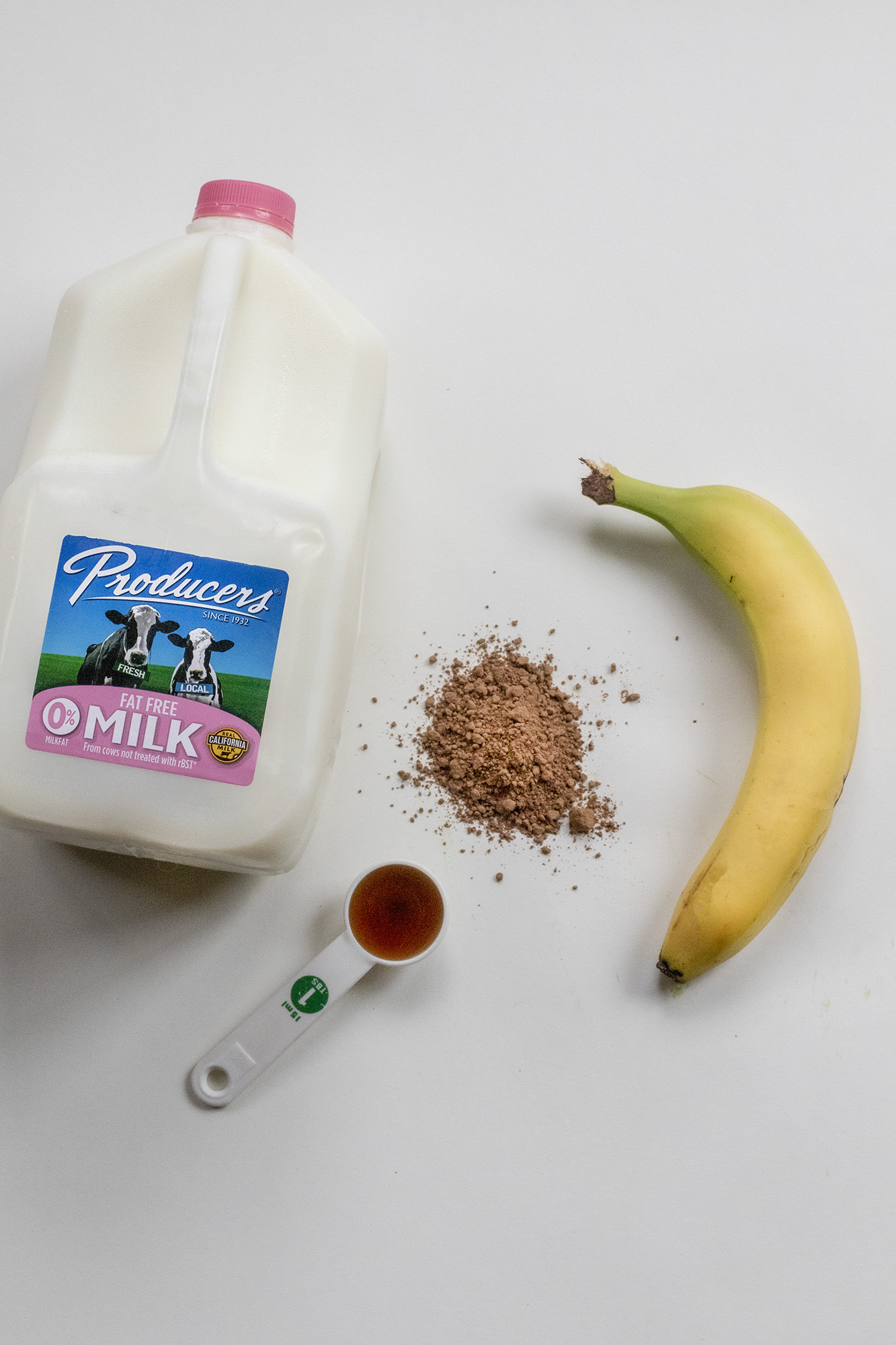 1Y8A7194 Producers Fat Free Gallon of milk, 1 tablespoon of maple syrup, cinnamon, and a banana laying on a flat white background.